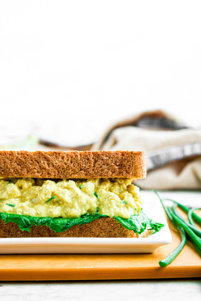 Head on view of a vegan egg salad sandwich on wheat bread with a lettuce leaf sitting on a wood cutting board