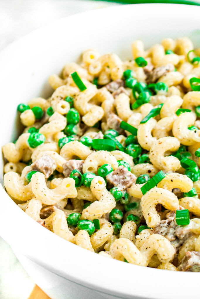 Close up overhead view of a round white bowl filled with creamy vegan pasta salad with diced seitan ham, peas, and sliced scallions