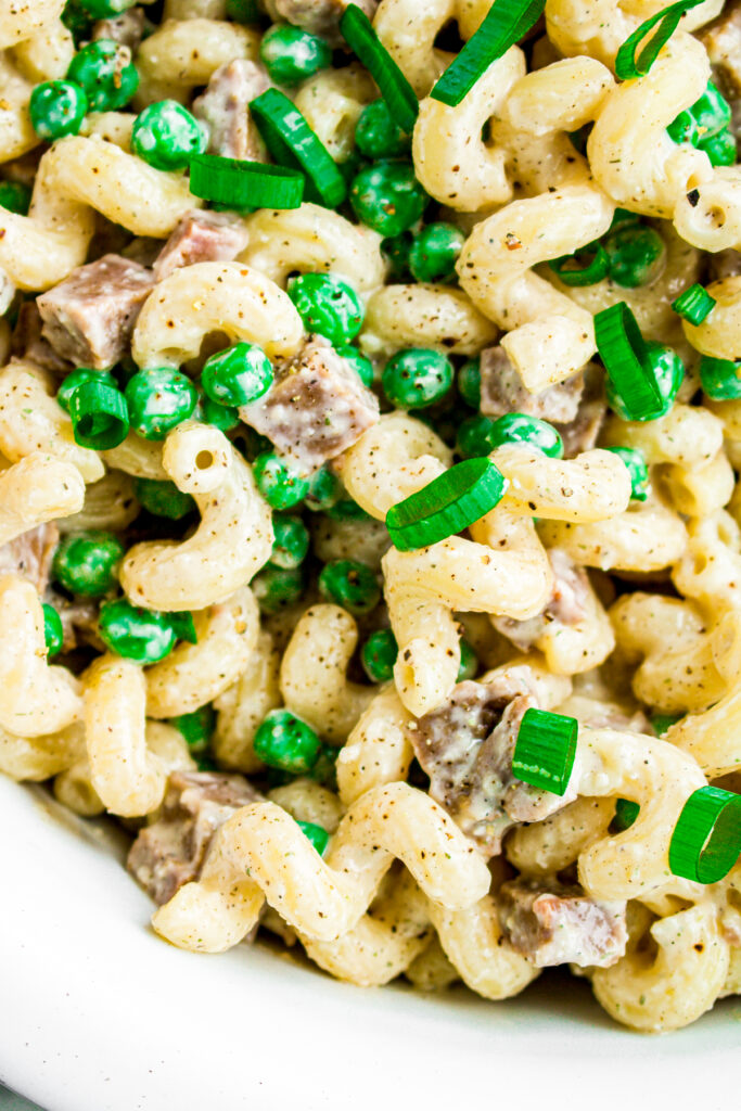Close up overhead view of a round white bowl filled with creamy vegan pasta salad with diced seitan ham, peas, and sliced scallions