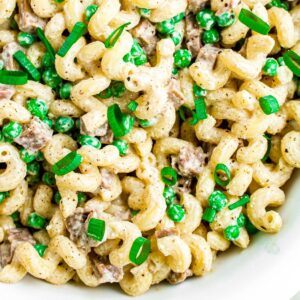 Overhead close up view of a round white bowl filled with creamy vegan pasta salad with diced seitan ham, peas, and sliced scallions