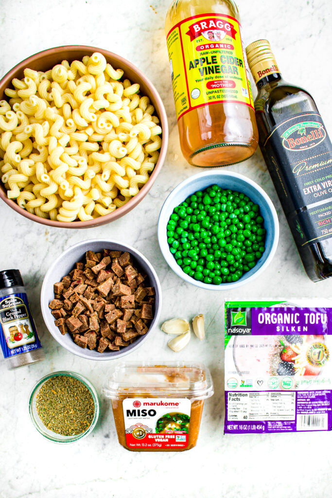 Overhead view of various ingredients laid out on a grey marble countertop like a bowl of cooked pasta, one with diced vegan ham, one with peas, bottles of apple cider vinegar and olive oil, a block of silken tofu, and a package of miso paste