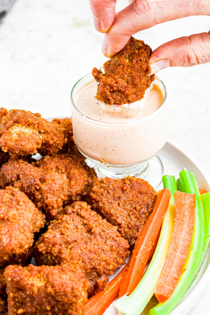 Overhead view of a white hand dipping a BBQ tofu wing into a creamy vegan bbq ranch dipping sauce