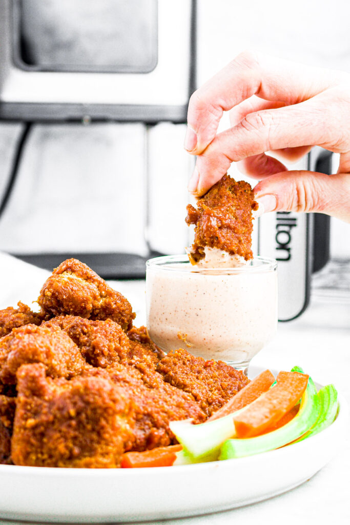 Overhead view of a white hand dipping a vegan BBQ wing into a creamy plant-based bbq ranch dipping sauce