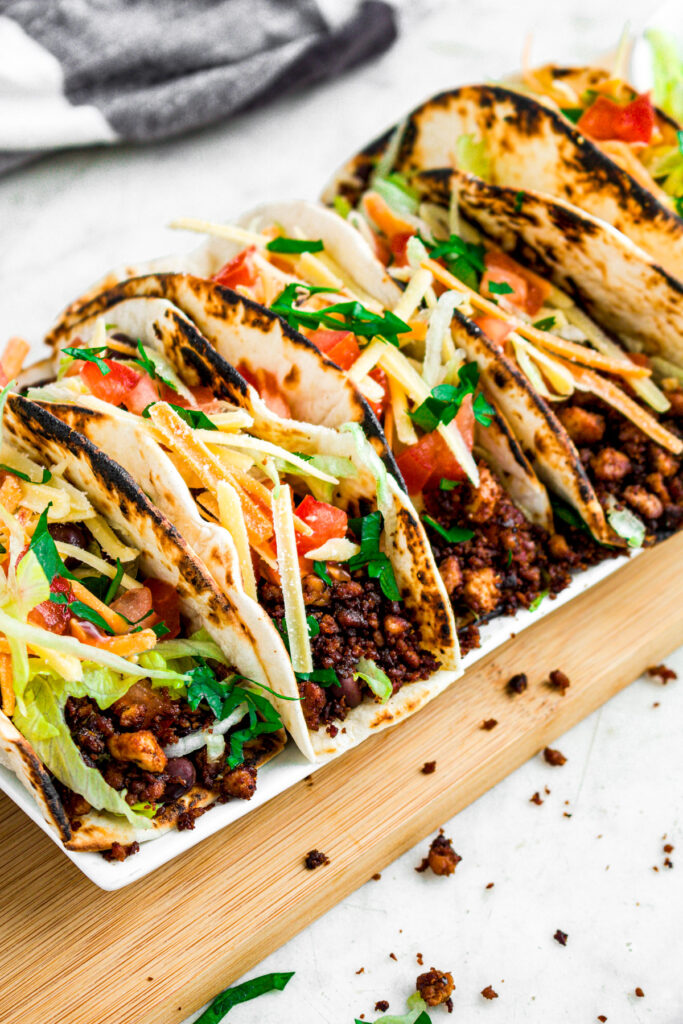 Overhead view of five tofu tacos topped with shredded lettuce, vegan cheese, tomatoes, and black beans on a long rectangular white plate