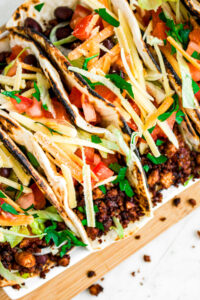 Overhead close up view of five tofu and black bean tacos topped with shredded lettuce, vegan cheese, and tomatoes on a long rectangular wood board
