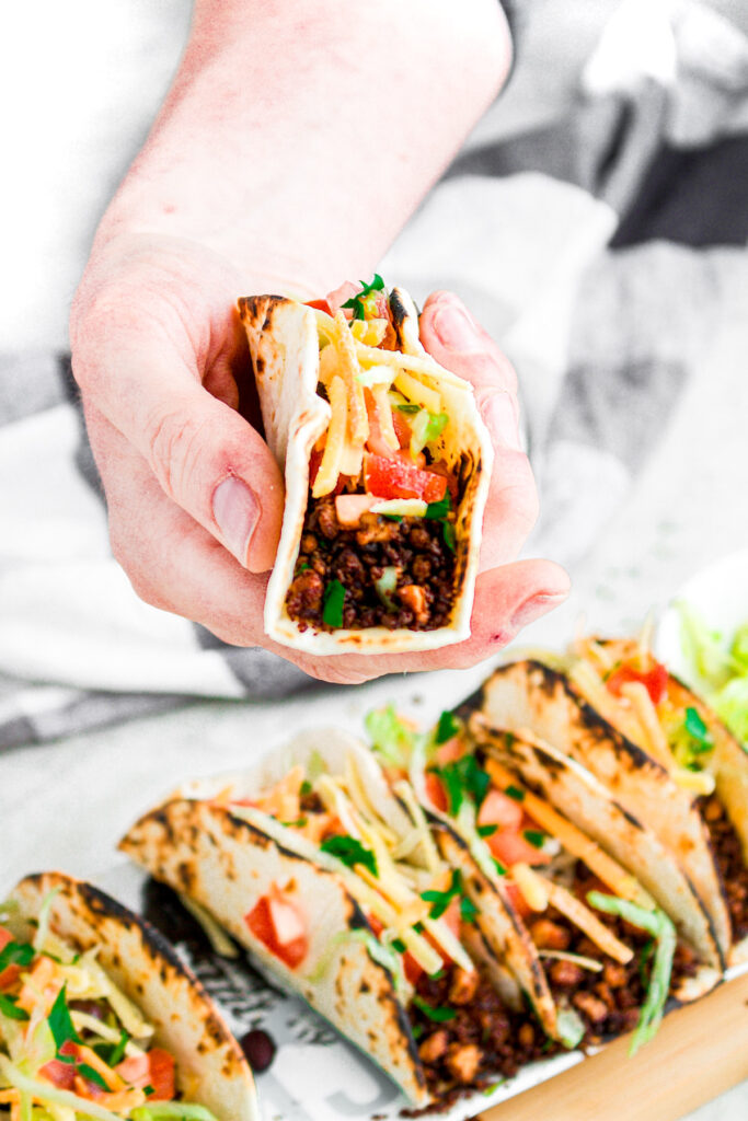 Head on view of a hand holding a vegan taco stuffed with tofu taco crumbles, diced tomatoes, shredded lettuce, and vegan cheese