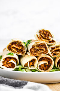 Head on view of eight vegan tofu taquitos stacked in a pyramid position on a round white plate on top of a bed of shredded green lettuce