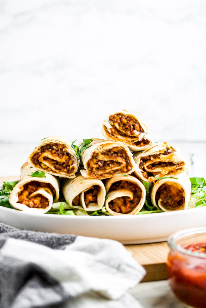 Head on view of eight vegan flautas stacked in a pyramid position on a round white plate on top of a bed of shredded green lettuce