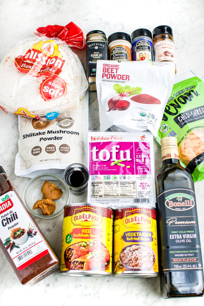 Overhead view of ingredients arranged on a grey marble backdrop including extra firm tofu, enchilada sauce, refried beans, spices, vegan cheddar, olive oil, tortillas, mushroom powder, and beet powder