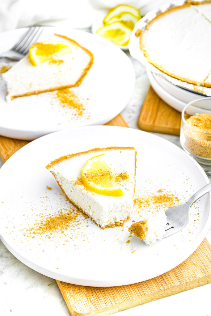Overhead view of a round white plate with a slice of creamy vegan lemon pie topped with a slice of lemon and graham cracker crumbs