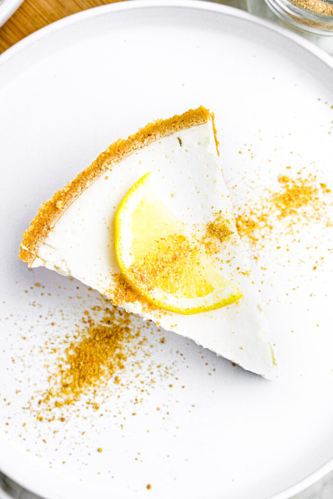 Overhead view of a round white plate with a slice of creamy vegan lemon pie topped with a slice of lemon and graham cracker crumbs