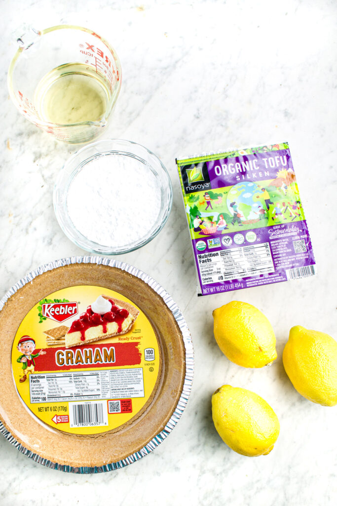 Overhead view of various ingredients laid out on a marble countertop including graham cracker crust, silken tofu, three lemons, powdered sugar, and coconut oil