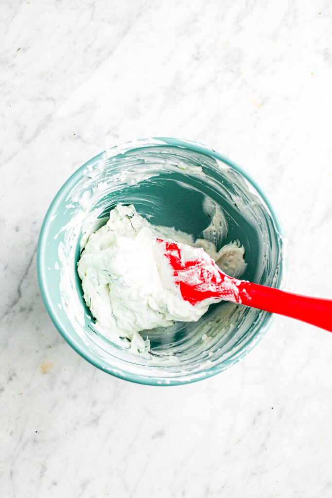 Overhead view of a small blue bowl filled with flavored vegan dill pickle cream cheese with fresh herbs being stirred by a red rubber spatula