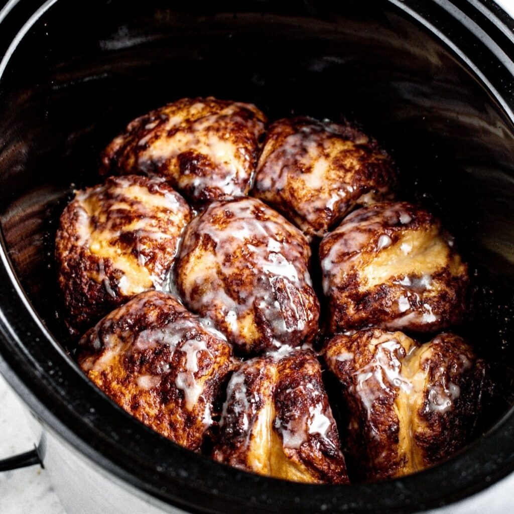 Overhead view of an oblong black and silver slow-cooker filled with crockpot vegan cinnamon roll casserole topped with dairy-free glaze