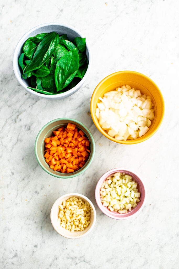 Overhead view of five bowls holding various chopped veggies like basil, onions, carrots, garlic, and celery
