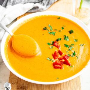 Overhead close up view of a round white bowl filled with creamy vegan red pepper soup topped with minced herbs and chopped red peppers