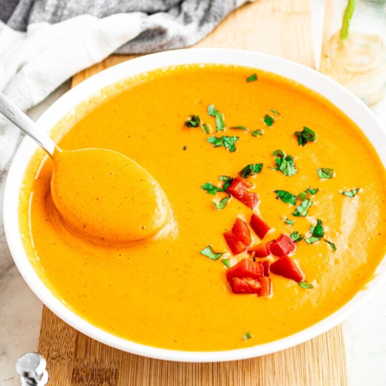 Overhead close up view of a round white bowl filled with creamy vegan red pepper soup topped with minced herbs and chopped red peppers