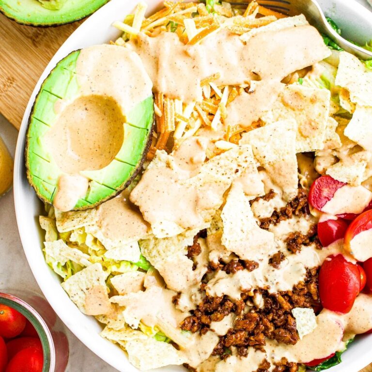 Overhead view of a round white high walled plate filled with vegan taco salad with walnut taco meat, half an avocado, cherry tomatoes, a sprinkle of tortilla chips, and a drizzle of cheesy taco salad dressing