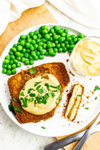 Overhead view of a round white plate with a rectangle of crispy baked tofu cordon bleu topped with creamy dijon sauce next to a pile of peas
