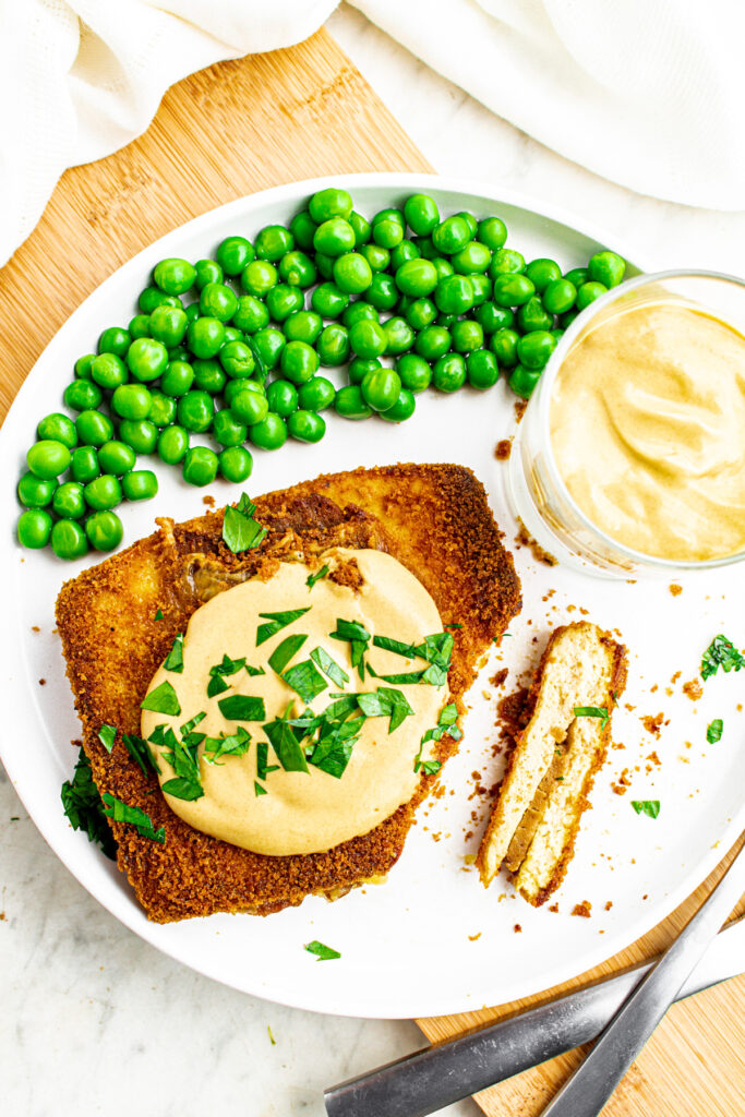 Overhead view of a round white plate with a rectangle of crispy baked tofu cordon bleu topped with creamy dijon sauce next to a pile of peas