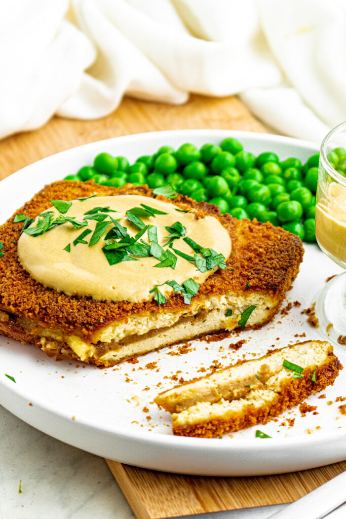 Overhead view of a round white plate with a rectangular breaded and baked vegan cordon bleu partially sliced next to a pile of green peas and a small clear bowl of dijon sauce
