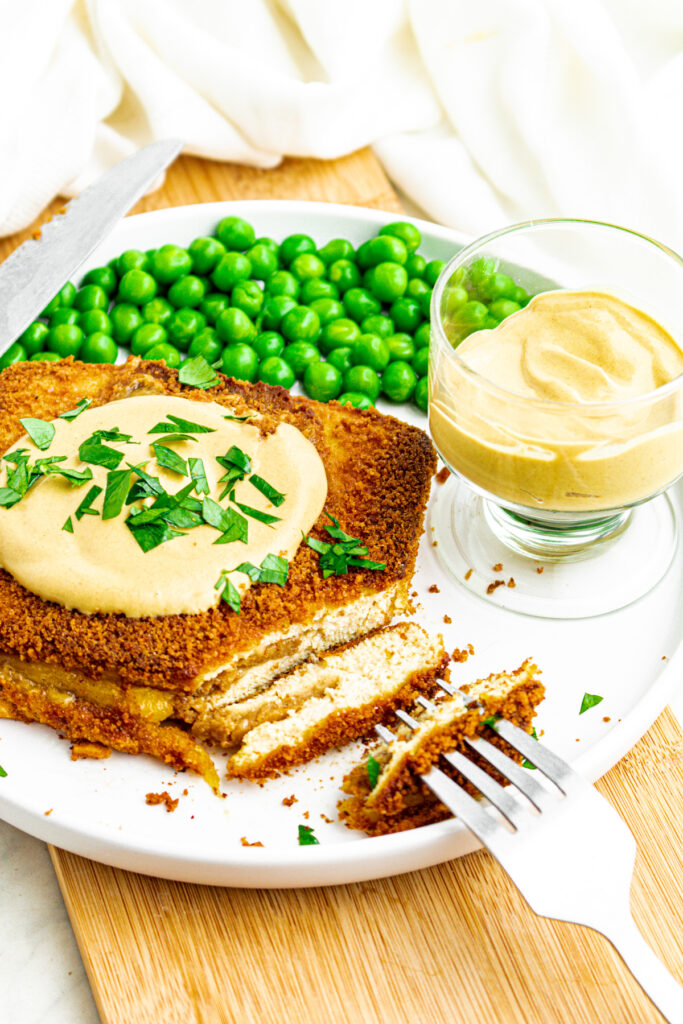 Overhead view of a round white plate with a rectangular breaded and baked vegan cordon bleu partially sliced next to a pile of green peas and a small clear bowl of dijon sauce
