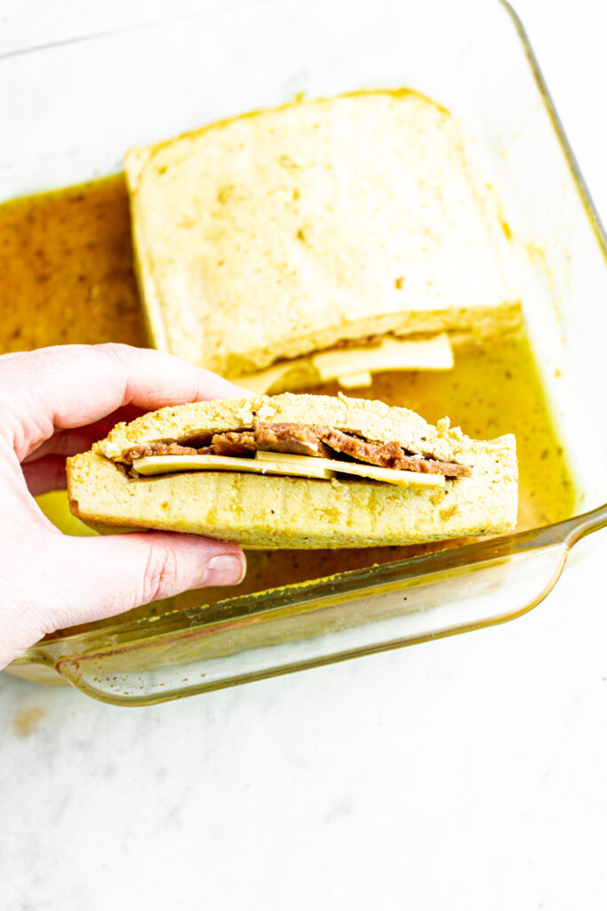 Overhead view of a hand holding a thick slice of tofu stuffed with seitan ham and vegan cheese slices