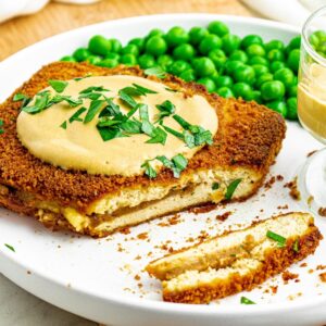 Overhead view of a round white plate with a rectangular breaded and baked vegan cordon bleu partially sliced next to a pile of green peas and a small clear bowl of dijon sauce