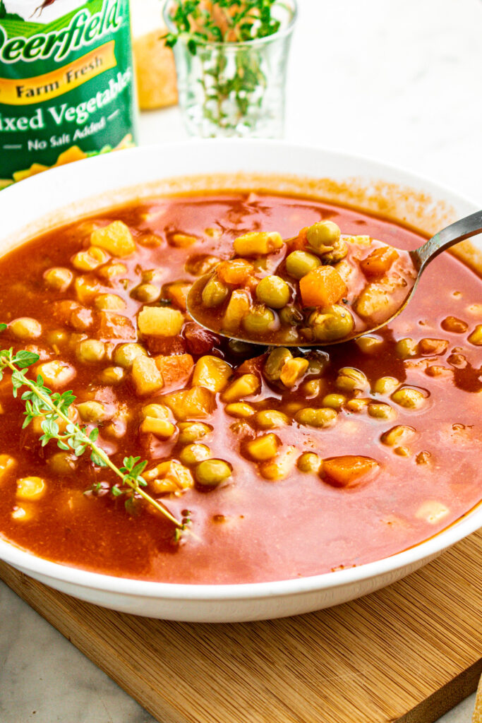 Overhead view of a round white bowl filled with v8 vegetable soup with a sprig of thyme on one side and a spoon digging into the other side