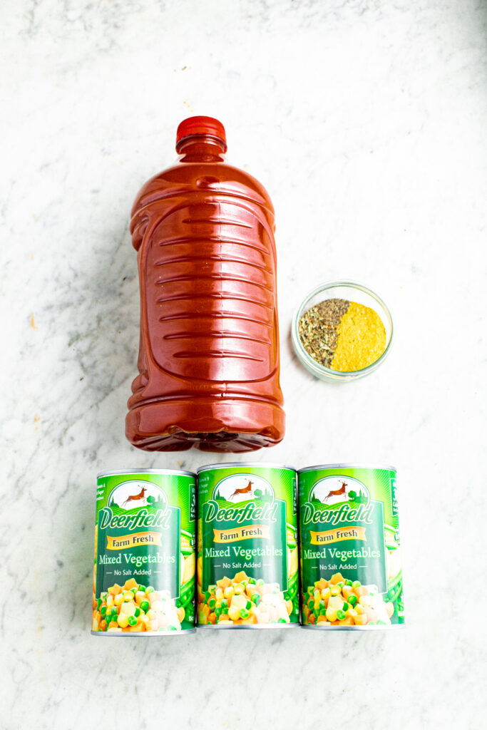 Overhead view of a large bottle of v8 juice, a clear glass bowl filled with spices, and three Deerfield mixed vegetables cans