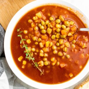 Overhead view of a round white bowl filled with v8 vegetable soup with a sprig of thyme on one side and a spoon digging into the other side