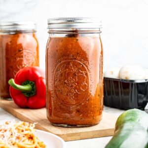 Head on view of a large glass jar filled with vegetable sauce for pasta surrounded by various vegetables