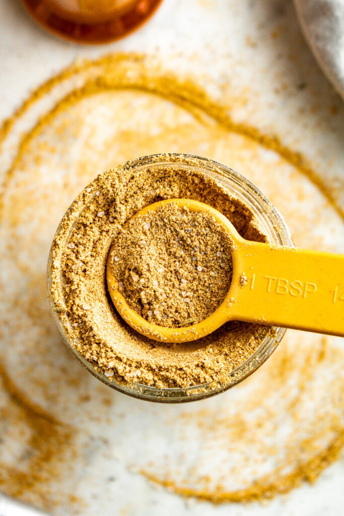 Overhead view of a clear glass jar filled with homemade vegan beef bouillon powder with a Tbsp digging into it and broth powder spilling over the edges