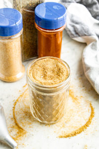 Overhead view of a clear glass jar filled with homemade vegan beef bouillon powder with spice jars behind it and broth powder spilling over the edges