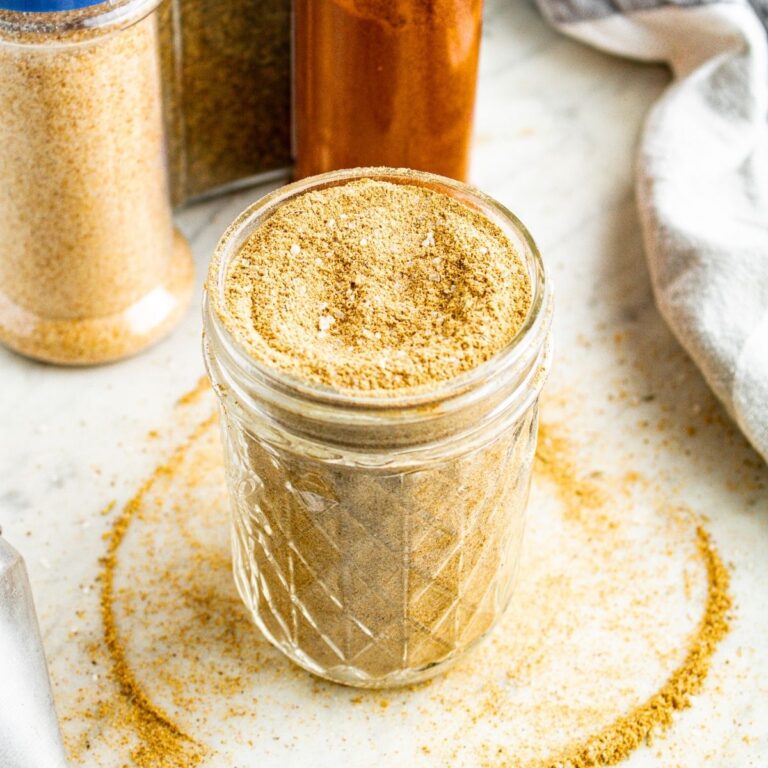 Overhead view of a clear glass jar filled with homemade vegan beef bouillon powder with spice jars behind it and broth powder spilling over the edges