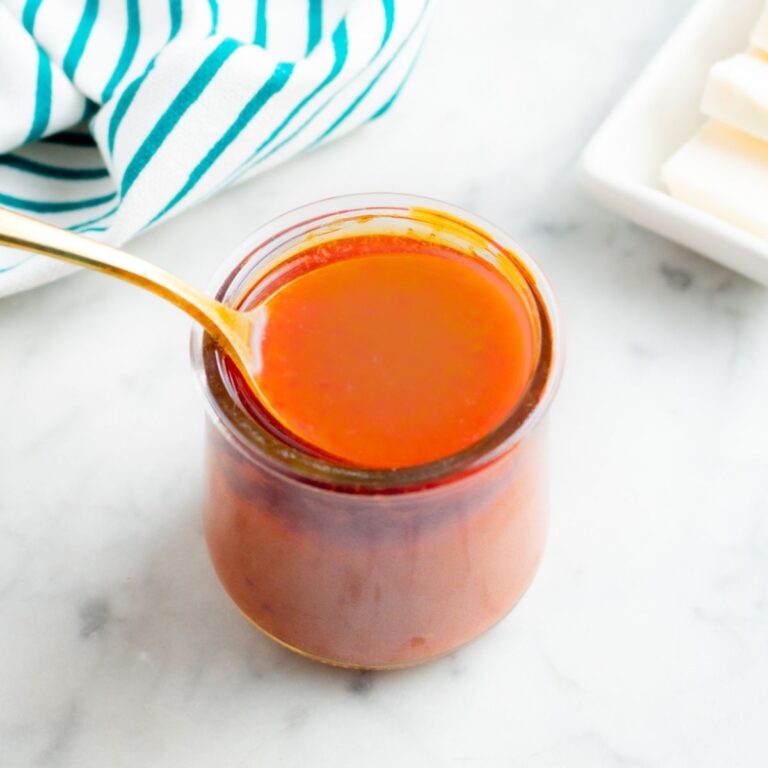 Overhead view of a clear glass jar filled with creamy vegan buffalo sauce with a gold spoon digging into it