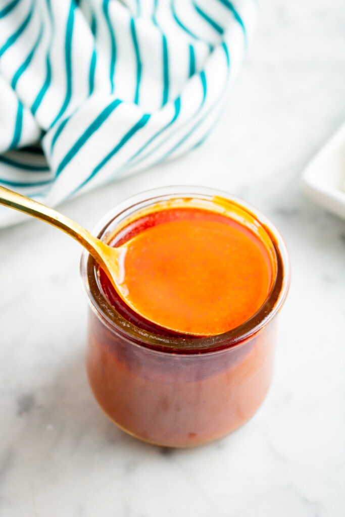 Overhead view of a clear glass jar filled with creamy vegan buffalo sauce with a gold spoon digging into it