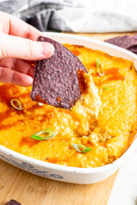 Overhead close up view of a blue tortilla chip being pulled out of a square dish of vegan buffalo chicken dip with a cheese pull