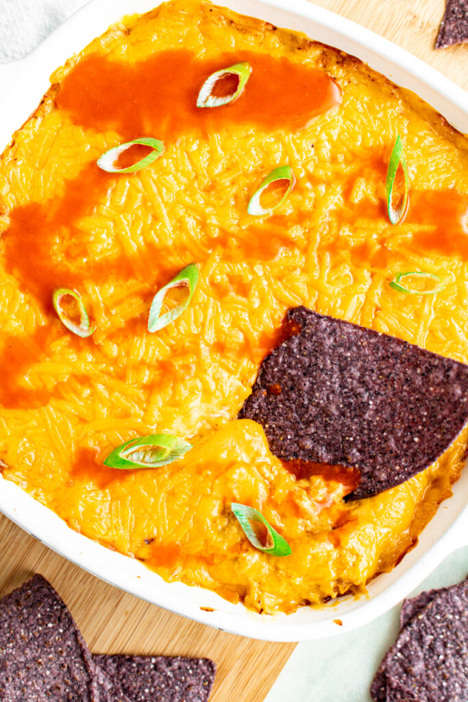 Overhead view of a square white baking dish of vegan buffalo chicken dip topped with sliced scallions and drizzled with hot sauce
