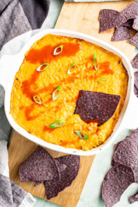 Overhead view of a square white baking dish of vegan buffalo chicken dip topped with sliced scallions and drizzled with hot sauce