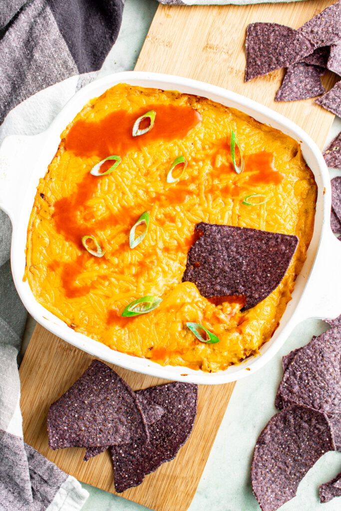 Overhead view of a square white baking dish of vegan buffalo chicken dip topped with sliced scallions and drizzled with hot sauce