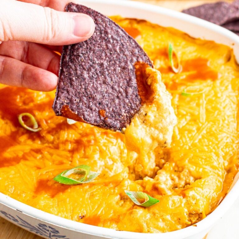 Overhead close up view of a blue tortilla chip being pulled out of a square dish of vegan buffalo chicken dip with a cheese pull
