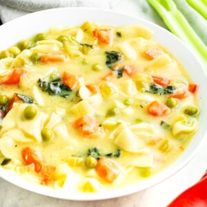 Overhead view of a round white bowl filled with vegetable noodle soup with celery behind the bowl and a red bell pepper in front of it