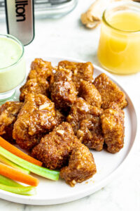 Overhead view of a round white plate piled high with vegan glazed sticky sweet wings with pineapple juice and fresh ginger in the background