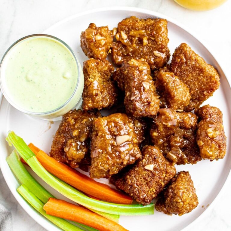 Overhead view of a round white plate with a pile of vegan pineapple wings on one side and carrot and celery sticks on the other with creamy avocado dip in the middle