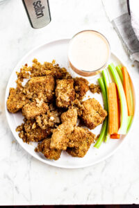 Overhead view of a round white plate with a pile of vegan garlic parmesan wings with a clear glass bowl of buffalo ranch and veggie sticks