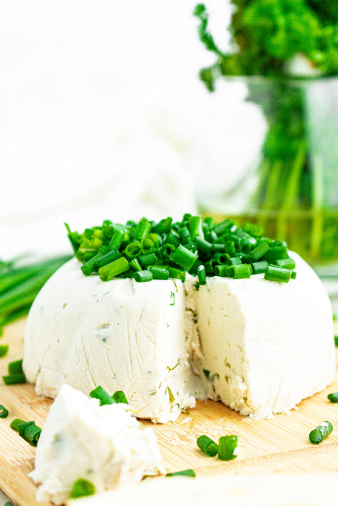 Close up head on view of a round wheel of creamy vegan Boursin cheese topped with sliced chives on a wood cutting board with a big chunk taken out