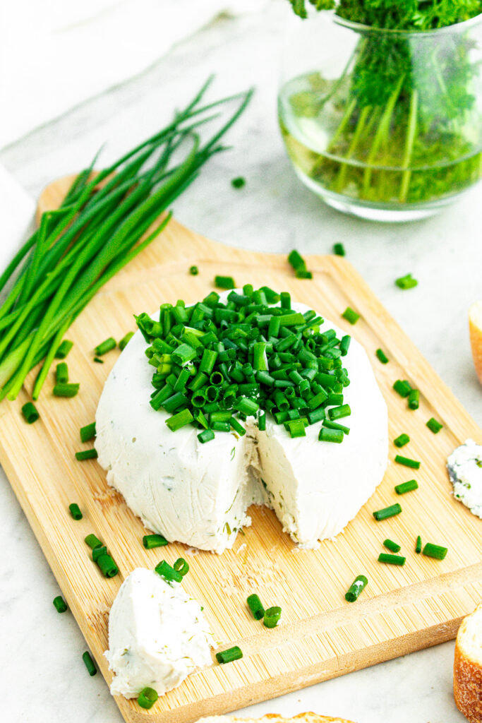 Overhead view of a round wheel of creamy vegan Boursin cheese topped with sliced chives on a wood cutting board with a big chunk taken out