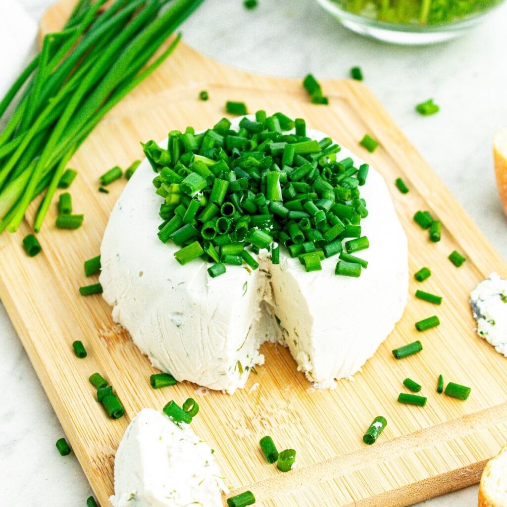 Overhead view of a round wheel of creamy vegan Boursin cheese topped with sliced chives on a wood cutting board