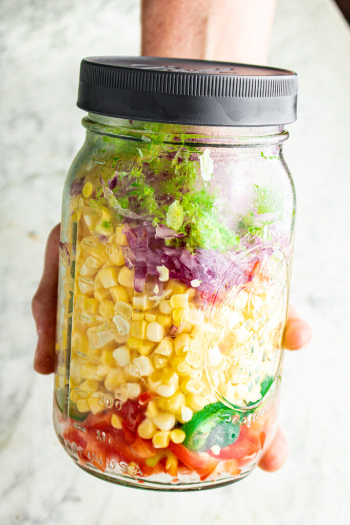 An outstreatched arm holding a clear glass jar full of chopped veggies like corn, red pepper, red onion, lime zest, and sliced jalapenos